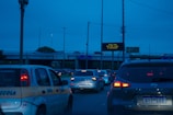 Traffic congestion on a road during the evening with numerous vehicles halted. A visible overhead sign displays traffic information while cars wait in line. The sky is a deep blue, suggesting an impending nightfall.
