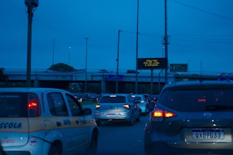 Traffic congestion on a road during the evening with numerous vehicles halted. A visible overhead sign displays traffic information while cars wait in line. The sky is a deep blue, suggesting an impending nightfall.
