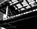 A black and white image depicting a partially constructed building with visible steel beams and girders. A construction worker is sitting on the edge of the structure, emphasizing the industrial and urban setting. The angles of the beams create a geometric pattern, and there are shadows cast on different surfaces.