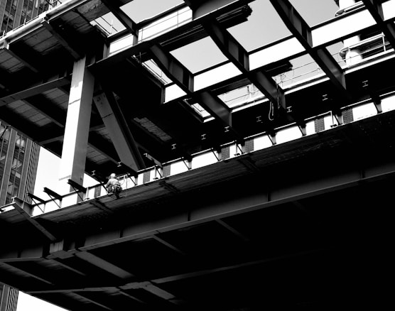 A black and white image depicting a partially constructed building with visible steel beams and girders. A construction worker is sitting on the edge of the structure, emphasizing the industrial and urban setting. The angles of the beams create a geometric pattern, and there are shadows cast on different surfaces.