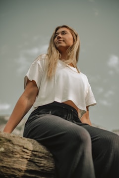 A person is seated on a wooden surface, gazing upward with a serene expression. The individual has long blonde hair and is wearing a white cropped t-shirt and dark pants. The background is a clear sky with a few clouds, giving a sense of calm and openness.