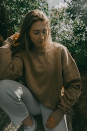 A young woman with long hair sits outdoors in a natural setting, surrounded by greenery. She is wearing a light brown sweatshirt and white pants, with one hand touching her hair.
