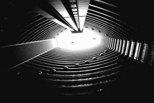 A dramatic black and white perspective of the inside of a tall circular building with multiple floors. The view is directed upwards towards the bright opening at the top, surrounded by concentric balconies that give an impression of depth and height.