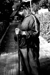 An elderly person stands on a paved path holding a walking stick. They wear a baseball cap and a jacket with stripes on the sleeves. The background features leafy bushes and another person walking away in the distance, creating depth in the scene.