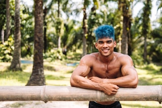 A muscular man with blue hair is smiling and leaning on a bamboo fence in a lush, sunlit forest with tall palm trees and a vibrant green landscape.