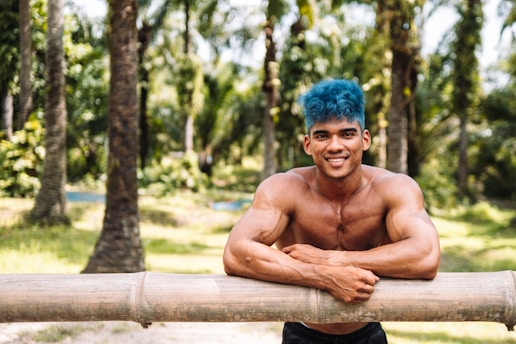A muscular man with blue hair is smiling and leaning on a bamboo fence in a lush, sunlit forest with tall palm trees and a vibrant green landscape.