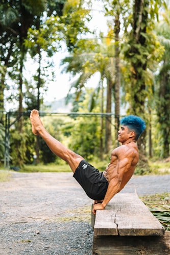 A muscular man with blue hair is performing a core exercise on a wooden bench outdoors. He is balancing his body with legs extended and feet off the ground in a lush green environment.