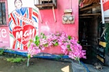 A mural on a pink wall features an illustration of a person with the words 'Art Capital' across their chest and 'Serbisyong May Puso' inside a heart. Pink flowers are arranged at the base of the wall, and there are two electrical meters above. Part of an open doorway is visible to the right.