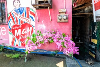 A mural on a pink wall features an illustration of a person with the words 'Art Capital' across their chest and 'Serbisyong May Puso' inside a heart. Pink flowers are arranged at the base of the wall, and there are two electrical meters above. Part of an open doorway is visible to the right.