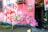 A mural on a pink wall features an illustration of a person with the words 'Art Capital' across their chest and 'Serbisyong May Puso' inside a heart. Pink flowers are arranged at the base of the wall, and there are two electrical meters above. Part of an open doorway is visible to the right.