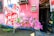A mural on a pink wall features an illustration of a person with the words 'Art Capital' across their chest and 'Serbisyong May Puso' inside a heart. Pink flowers are arranged at the base of the wall, and there are two electrical meters above. Part of an open doorway is visible to the right.