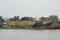 Several large boats under construction are positioned by a riverside. The hulls are surrounded by scaffolding and wooden supports. Yellow and reddish paint is visible on parts of the structures. In the background, there are trees, small buildings, and power lines under an overcast sky.