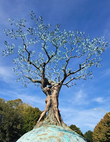 A bronze sculpture depicting two human figures intertwining and transforming into the trunk and branches of a tree. The tree's leaves are metallic, shimmering in the sunlight against a clear blue sky. The sculpture is set against a backdrop of natural foliage, suggesting a blend of nature and art.