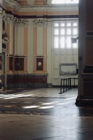 An elegant interior space with high ceilings and ornate architectural details. The walls feature intricate molding and columns with decorative elements. Large windows allow natural light to flood the space, creating patterns on the floor. A neatly organized information board is visible near the windows, and there are barriers set up likely for directing foot traffic.