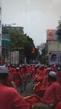 A large group of people in matching red outfits and white hats are marching down a street, carrying drums. Some are playing the drums while others hold flags, and they are surrounded by city buildings. A banner featuring a face is visible on one of the buildings. Trees and scaffolding are also seen along the street.