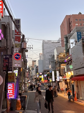 A bustling city street scene with numerous signs written in Korean, displaying a mix of bright neon lights and shopfronts. People are walking along the street, suggesting a lively atmosphere. Buildings of different heights line both sides of the street, indicating an urban setting during early evening.