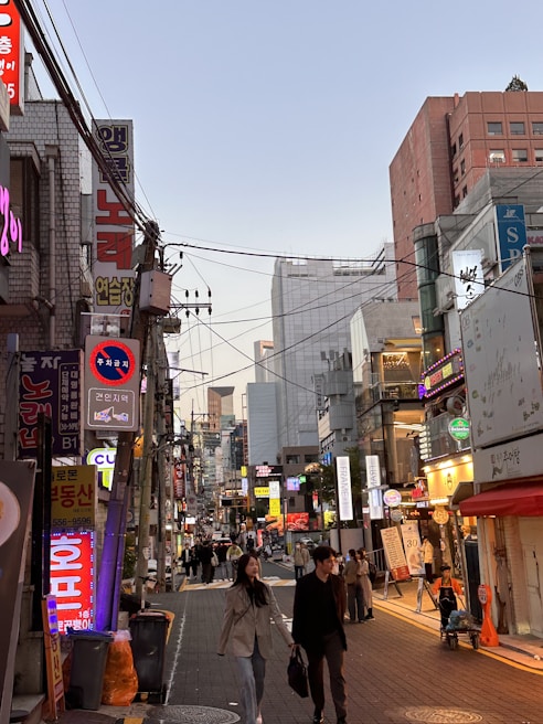 A bustling city street scene with numerous signs written in Korean, displaying a mix of bright neon lights and shopfronts. People are walking along the street, suggesting a lively atmosphere. Buildings of different heights line both sides of the street, indicating an urban setting during early evening.