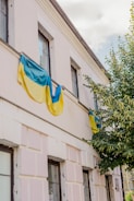 A building facade partially covered by Ukrainian flags, with a tree next to it. The building has multiple windows and is painted in a light color, possibly white or cream. The sky is visible above, partially cloudy.