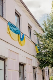 A building facade partially covered by Ukrainian flags, with a tree next to it. The building has multiple windows and is painted in a light color, possibly white or cream. The sky is visible above, partially cloudy.