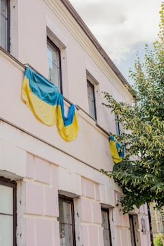 A building facade partially covered by Ukrainian flags, with a tree next to it. The building has multiple windows and is painted in a light color, possibly white or cream. The sky is visible above, partially cloudy.