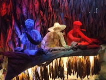 Three wireframe figures are seated behind a table, each colored differently in vivid blue, yellow, and red hues. They appear to be engaged in the process of rolling cigars, with numerous cigars placed in front of them. The background is filled with hanging tobacco leaves, creating a dynamic and artistic atmosphere.