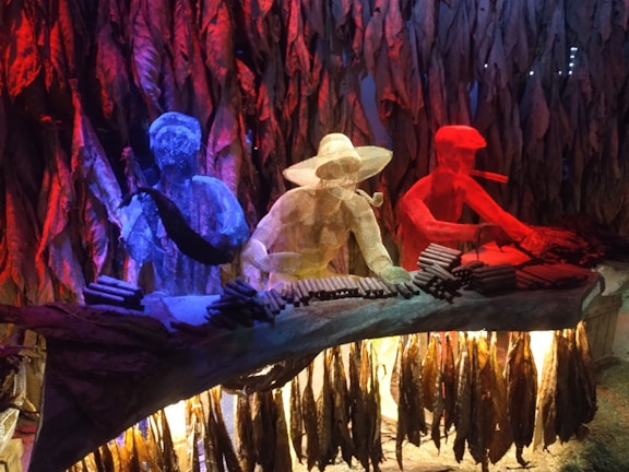 Three wireframe figures are seated behind a table, each colored differently in vivid blue, yellow, and red hues. They appear to be engaged in the process of rolling cigars, with numerous cigars placed in front of them. The background is filled with hanging tobacco leaves, creating a dynamic and artistic atmosphere.