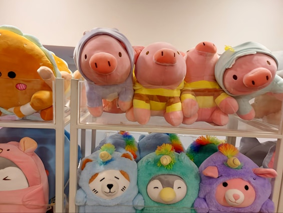 A collection of colorful plush toys on display. The upper shelf holds pig plushies dressed in various outfits, including striped yellow shirts and hoodies. The lower shelf features plush toys resembling animals in pastel hues, such as a blue bear, a penguin, and a purple llama.