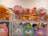 A collection of colorful plush toys on display. The upper shelf holds pig plushies dressed in various outfits, including striped yellow shirts and hoodies. The lower shelf features plush toys resembling animals in pastel hues, such as a blue bear, a penguin, and a purple llama.