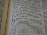 An open book with English text printed on two visible pages, featuring a section from a religious scripture with chapter and verse numbers clearly marked. The lighting casts a mild shadow across the pages, and the text is in black font on a cream-colored paper.