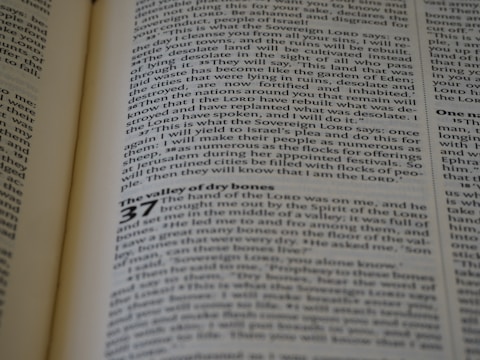 An open book with English text printed on two visible pages, featuring a section from a religious scripture with chapter and verse numbers clearly marked. The lighting casts a mild shadow across the pages, and the text is in black font on a cream-colored paper.