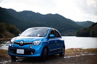 A blue Renault car is parked near a lake with lush, green hills in the background. The setting appears serene with a calm body of water, surrounded by dense forested hills under a partly cloudy sky.