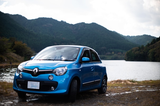 A blue Renault car is parked near a lake with lush, green hills in the background. The setting appears serene with a calm body of water, surrounded by dense forested hills under a partly cloudy sky.