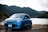 A blue Renault car is parked near a lake with lush, green hills in the background. The setting appears serene with a calm body of water, surrounded by dense forested hills under a partly cloudy sky.
