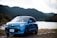 A blue Renault car is parked near a lake with lush, green hills in the background. The setting appears serene with a calm body of water, surrounded by dense forested hills under a partly cloudy sky.