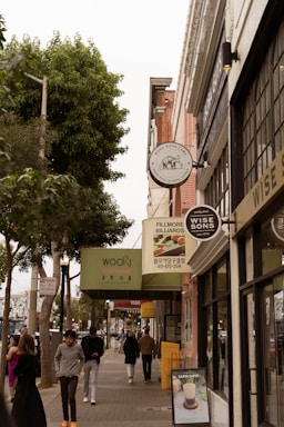 A busy urban street lined with various shops and restaurants, featuring several storefront signs including 'Fillmore Billiards' and 'Wise Sons'. People walk along the sidewalk, some carrying bags and others looking at their phones. There are tall trees on the left side, providing greenery amidst the city setting.