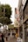 A busy urban street lined with various shops and restaurants, featuring several storefront signs including 'Fillmore Billiards' and 'Wise Sons'. People walk along the sidewalk, some carrying bags and others looking at their phones. There are tall trees on the left side, providing greenery amidst the city setting.