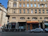 A multi-story historical building features ornate architectural details like brick and stonework. The ground floor has businesses with signage, including a cafe and a store with a closed entrance. Several windows are visible on the upper floors with decorative frames. There is a parked silver station wagon in front of the building, with a small outdoor seating area to one side.
