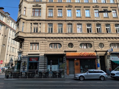 A multi-story historical building features ornate architectural details like brick and stonework. The ground floor has businesses with signage, including a cafe and a store with a closed entrance. Several windows are visible on the upper floors with decorative frames. There is a parked silver station wagon in front of the building, with a small outdoor seating area to one side.