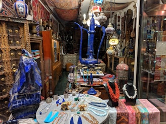 A richly decorated shop interior showcasing a variety of traditional and cultural items. In the foreground, an array of colorful necklaces is displayed on black busts alongside other jewelry pieces on a small table. The centerpiece is a large, ornate blue hookah. The background is filled with intricately designed lamps, vases, and textiles, all adding to the eclectic and vibrant atmosphere.
