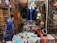 A richly decorated shop interior showcasing a variety of traditional and cultural items. In the foreground, an array of colorful necklaces is displayed on black busts alongside other jewelry pieces on a small table. The centerpiece is a large, ornate blue hookah. The background is filled with intricately designed lamps, vases, and textiles, all adding to the eclectic and vibrant atmosphere.