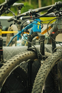 A close-up view of several mountain bikes mounted on a vehicle, featuring the bikes' handlebars and large, knobby tires. The main colors are in shades of blue, black, and yellow, and there is a background blur of a natural setting with green foliage.