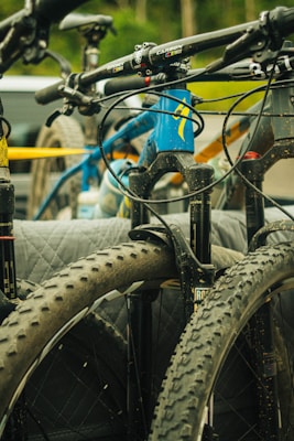A close-up view of several mountain bikes mounted on a vehicle, featuring the bikes' handlebars and large, knobby tires. The main colors are in shades of blue, black, and yellow, and there is a background blur of a natural setting with green foliage.