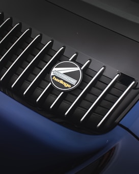 A close-up of a car's rear grille featuring the Porsche logo with the word 'heritage' written beneath it. The grille is composed of black vertical slats with silver accents.