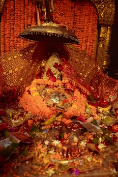 A beautifully adorned altar with a central deity figure covered in vibrant marigold garlands and red cloth, surrounded by various flowers and offerings. Intricate metal embellishments and patterns decorate the area, emanating a sense of reverence and devotion.