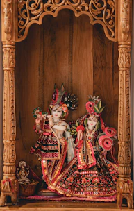 Two intricately decorated statues are placed within a wooden alcove featuring ornate carvings. The statues are adorned with vibrant traditional clothing and detailed embellishments, featuring patterns of red, pink, green, and gold. The expressive detailing on the faces and the elaborate headdresses give a lifelike presence. A smaller figure sits beside them, adding a contrasting scale.
