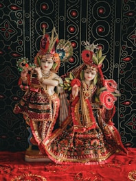 Two ornately dressed figures, resembling deities, adorned in richly decorated garments featuring vibrant patterns with intricate beadwork and embroidery. The figures are posed against a black backdrop featuring a symmetrical, circular pattern in red and white.