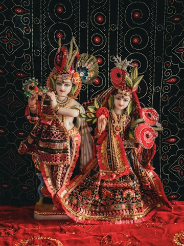 Two ornately dressed figures, resembling deities, adorned in richly decorated garments featuring vibrant patterns with intricate beadwork and embroidery. The figures are posed against a black backdrop featuring a symmetrical, circular pattern in red and white.