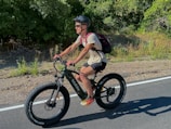 A person with a helmet and sunglasses rides a fat-tire bike along a paved road surrounded by greenery. They have a backpack and are wearing a light-colored shirt and dark shorts, with brightly colored shoes.