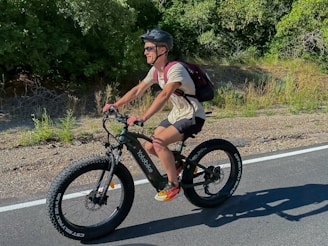 A person with a helmet and sunglasses rides a fat-tire bike along a paved road surrounded by greenery. They have a backpack and are wearing a light-colored shirt and dark shorts, with brightly colored shoes.
