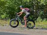 A person is riding a green mountain bike on a gravel path with large, thick tires. The individual is wearing a helmet, a red backpack, and athletic clothing. The background is filled with dense green foliage, suggesting a lush and natural environment.